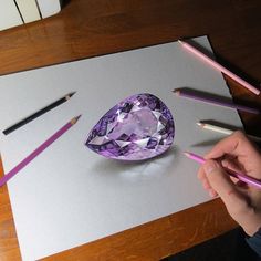 a person holding pencils near a purple diamond on a piece of paper with colored pencils next to it