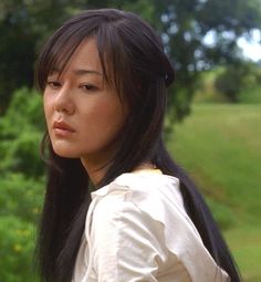 a woman with long black hair standing in front of green grass and trees, looking off to the side