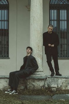 two men standing next to each other in front of a tall column with one man sitting on it