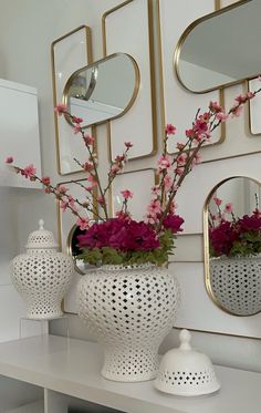 two white vases with pink flowers in them on a table next to mirrors and lamps