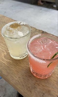 two cocktails sitting on top of a wooden table next to each other, one with a slice of lime