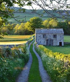 an old stone house sitting in the middle of a lush green field next to a dirt road