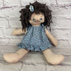 a stuffed doll with dreadlocks sitting against a white brick wall and wearing a blue dress