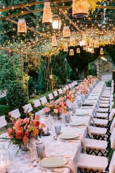 an outdoor dinner table set up with white linens and place settings, lit by hanging lights