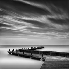 a black and white photo of a pier on the water with clouds in the sky