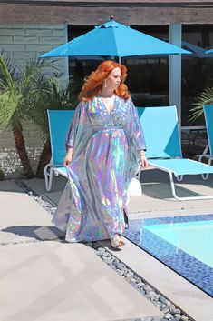 a woman standing next to a pool with an umbrella over her head and wearing a silver dress