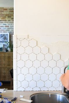 a woman standing in front of a kitchen sink next to a wall with hexagonal tiles on it