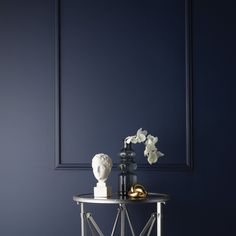 a white vase with flowers on top of a small table in front of a blue wall