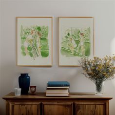 two paintings hang on the wall above a dresser with books and vases filled with flowers