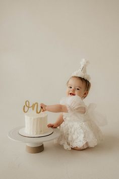 a baby girl in a white dress holding a cake with the number one on it