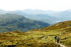 two people hiking up a grassy hill with mountains in the background