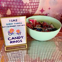 a bowl of candy sits next to a sign that says i like shiny things but i'd marry you with candy rings