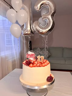 a birthday cake with white frosting and fruit on top, surrounded by silver balloons