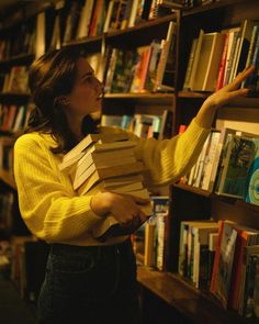 a woman is holding books in front of a book shelf full of books and looking at them