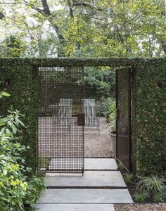 an open gate in the middle of a garden surrounded by trees and bushes, with two chairs on either side