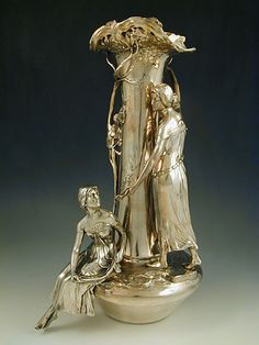 a silver vase with a woman and child sitting on the ground in front of it