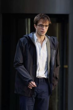 a man wearing glasses and a black jacket is standing in front of a building with his hands in his pockets