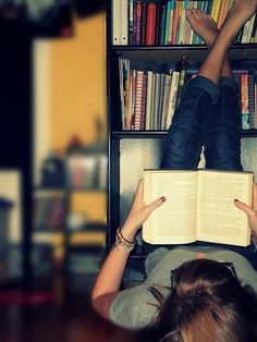 a woman laying down reading a book in front of a bookshelf filled with books