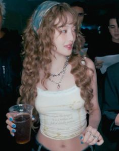 a woman with curly hair holding a drink in her right hand and wearing a white tank top