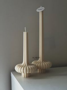 two tall white candles sitting on top of a table next to each other in front of a gray wall