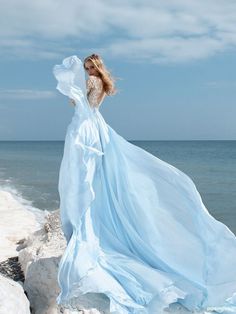 a woman in a long blue dress standing on rocks near the ocean with her hair blowing in the wind