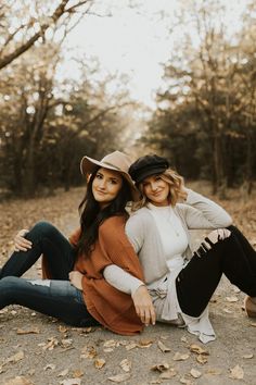 two women are sitting on the ground in front of some trees and leaves, one is wearing a hat