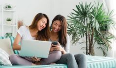 two young women sitting on a couch looking at a cell phone and laptop computer screen