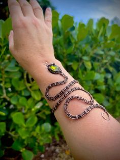 a woman's arm with a snake bracelet on top of it and green leaves in the background