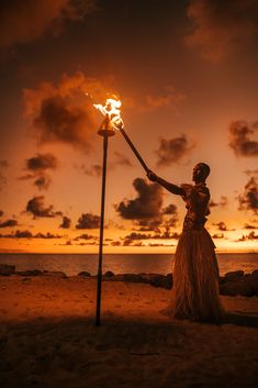 a woman holding a torch in her hand on the beach at sunset with clouds behind her