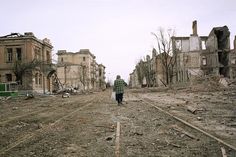 a man walking down a dirt road in front of buildings that have been burned out