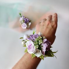 two wrist corsages with purple flowers and greenery on the wrist, one being held by someone's hand