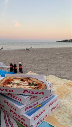 three pizza boxes stacked on top of each other near the beach with bottles of soda