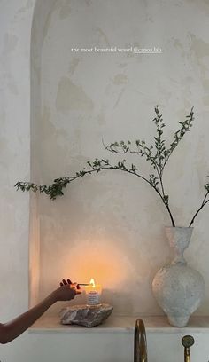 a person lighting a candle on a shelf next to a vase filled with greenery