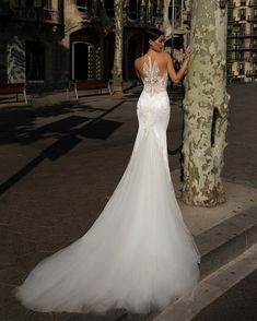 a woman in a white wedding dress standing next to a tree on the side walk