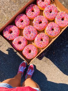 a box full of pink sprinkled donuts with one person's feet on the ground