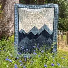 a blue and white quilt hanging from a tree in the grass with wildflowers