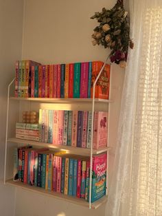 a book shelf filled with lots of books next to a white curtain and window sill