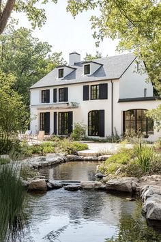 a white house with black shutters next to a stream and trees in the background