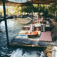 an outdoor seating area with hammocks and rugs on the water's edge