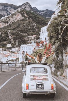 a woman standing on top of a car in the middle of a road with mountains behind her