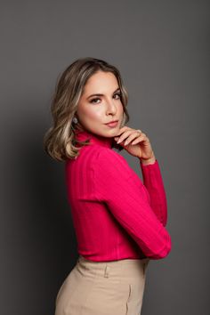 a woman in a pink sweater and tan pants poses for the camera with her hand on her chin