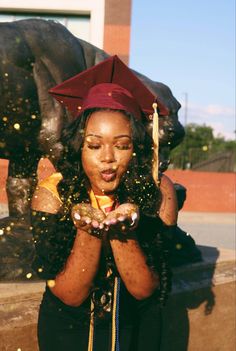 a woman wearing a graduation cap and gown blowing confetti in front of a statue