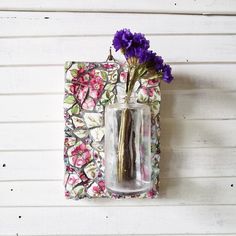 a vase with purple flowers in it sitting on a white wooden wall next to a flower pot