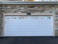 two white garage doors in front of a stone wall and black asphalt driveway with trees