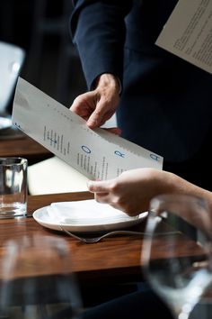 two people sitting at a table with plates and wine glasses on it, one holding a piece of paper