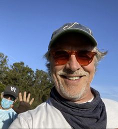 a man wearing sunglasses and a hat standing next to another man in front of trees