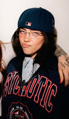 two women with tattoos and baseball caps posing for a photo together in front of a white wall