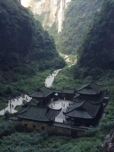 an old building in the middle of a valley surrounded by green trees and mountains with people walking around it