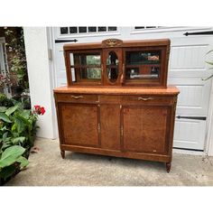 an old wooden china cabinet in front of a garage door with flowers and plants around it