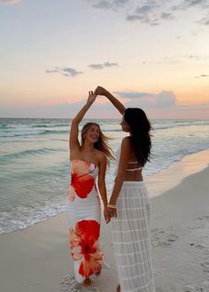 two beautiful women standing on top of a sandy beach next to the ocean at sunset